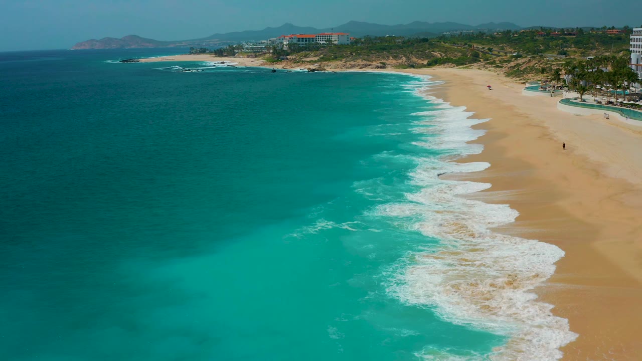 The aerial view holds on Secrets Marquis Los Cabos, Mexico, as a larger white foam band spreads across the lower right edge of the beach.