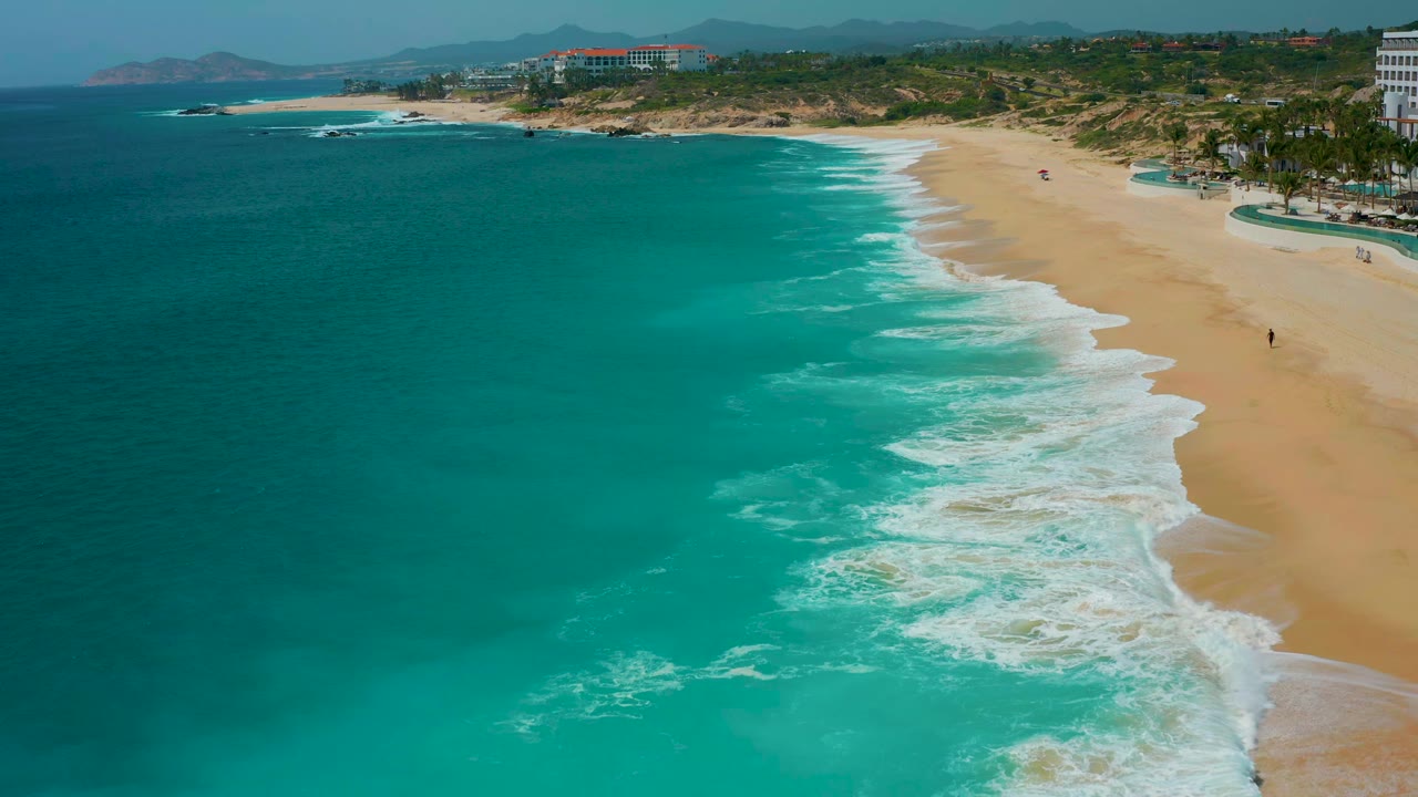 Aerial view at Secrets Marquis Los Cabos, Mexico, showing white wave bands curling diagonally along tan sand, with teal ocean filling most of the frame and no visible people.