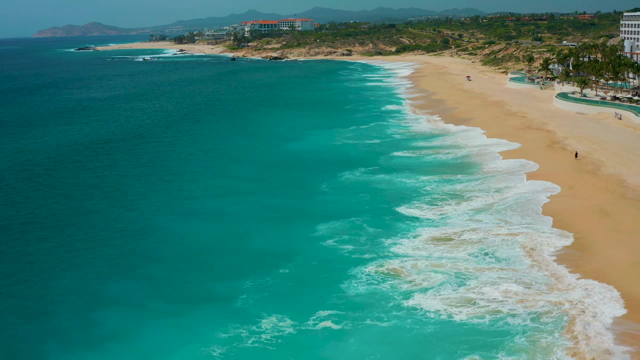 Drone-style frame over Secrets Marquis Los Cabos, Mexico, with foamy surf spreading farther across the lower beach, teal water on the left, shoreline development above, and no visible people.
