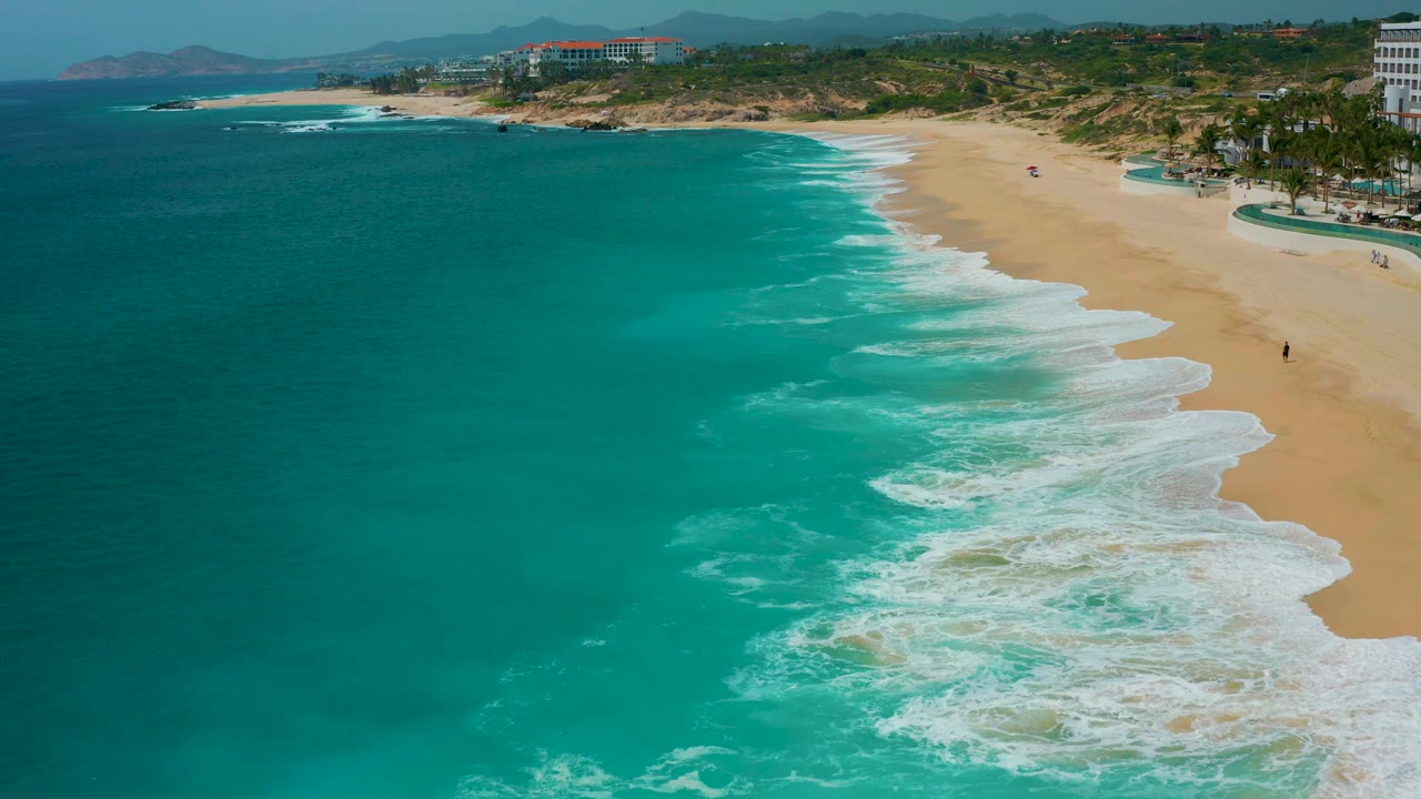 Oblique aerial frame at Secrets Marquis Los Cabos, Mexico, with deep teal water at left, a broad empty beach at right, thin white surf lines, and no visible people.