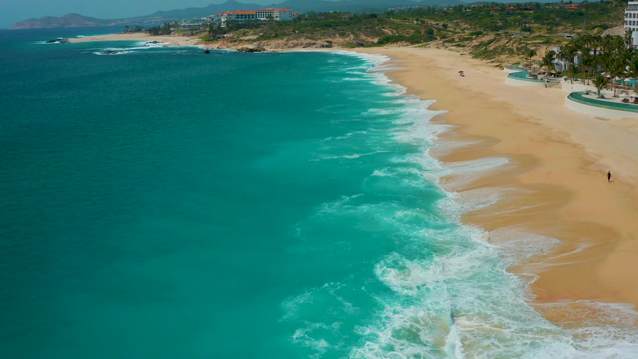 Aerial view at Secrets Marquis Los Cabos, Mexico, with turquoise water on the left, white surf crossing the sand, green coastal land beyond the beach, and no visible people.