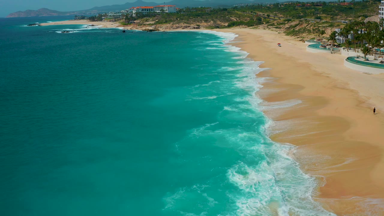 At 0:02, the beach curves along Secrets Marquis Los Cabos, Mexico, with active white surf in the center, clear water to the left, and resort structures above the sand with no visible people.