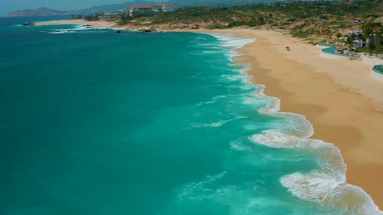 High aerial view at Secrets Marquis Los Cabos, Mexico, with turquoise water at left, white surf lines, tan beach at right, resort buildings beyond the bluff, and tiny beach users visible.