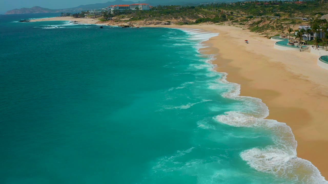Aerial frame over Secrets Marquis Los Cabos, Mexico, showing blue-green ocean covering most of the image, curved foam bands on the shoreline, pale sand, and small beach activity on the right.