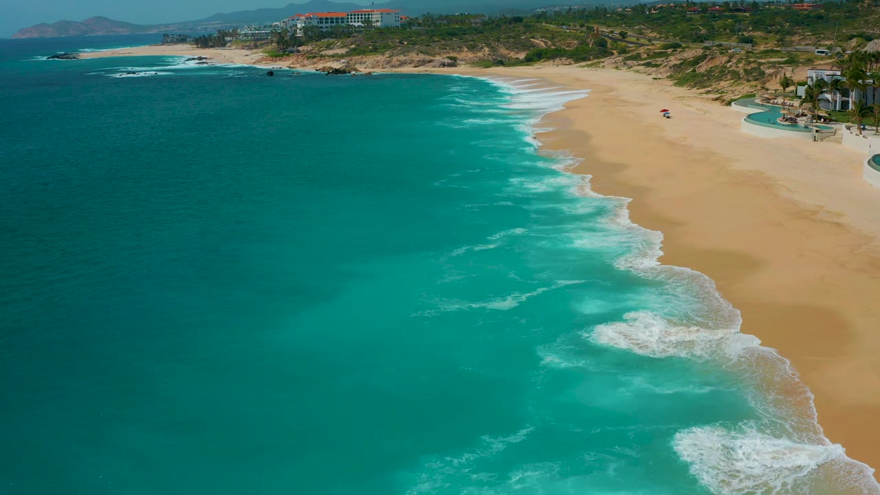 Oblique aerial view at Secrets Marquis Los Cabos, Mexico, with open turquoise water in the foreground, diagonal white surf, tan beach, low bluffs, resort structures, and no close-up faces.