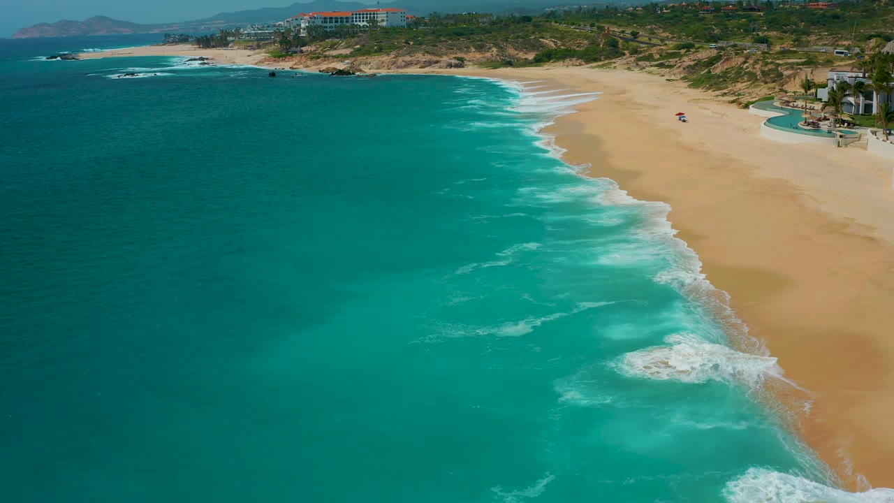 Wide aerial beach view with turquoise sea and resort buildings in the distance.