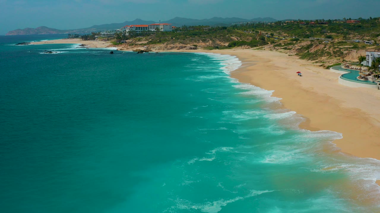 Opening aerial view at Secrets Marquis Los Cabos, Mexico, with turquoise water on the left, tan beach on the right, distant hills and low buildings beyond the shore, and no visible people.