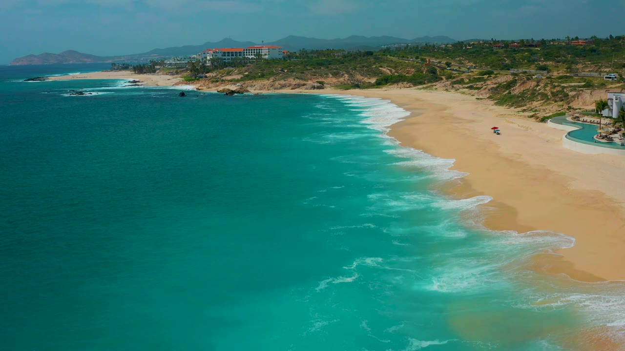 The view shifts along the Secrets Marquis Los Cabos shoreline, showing more open water in front, a long sweep of sand, rocky points to the left, and no visible people on the beach.