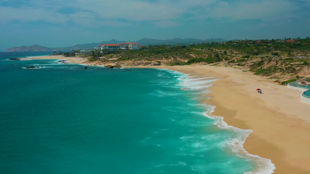 Aerial view at Secrets Marquis Los Cabos, Mexico, with teal ocean meeting a wide sandy beach, low dunes behind it, distant buildings near the coast, and no visible people.