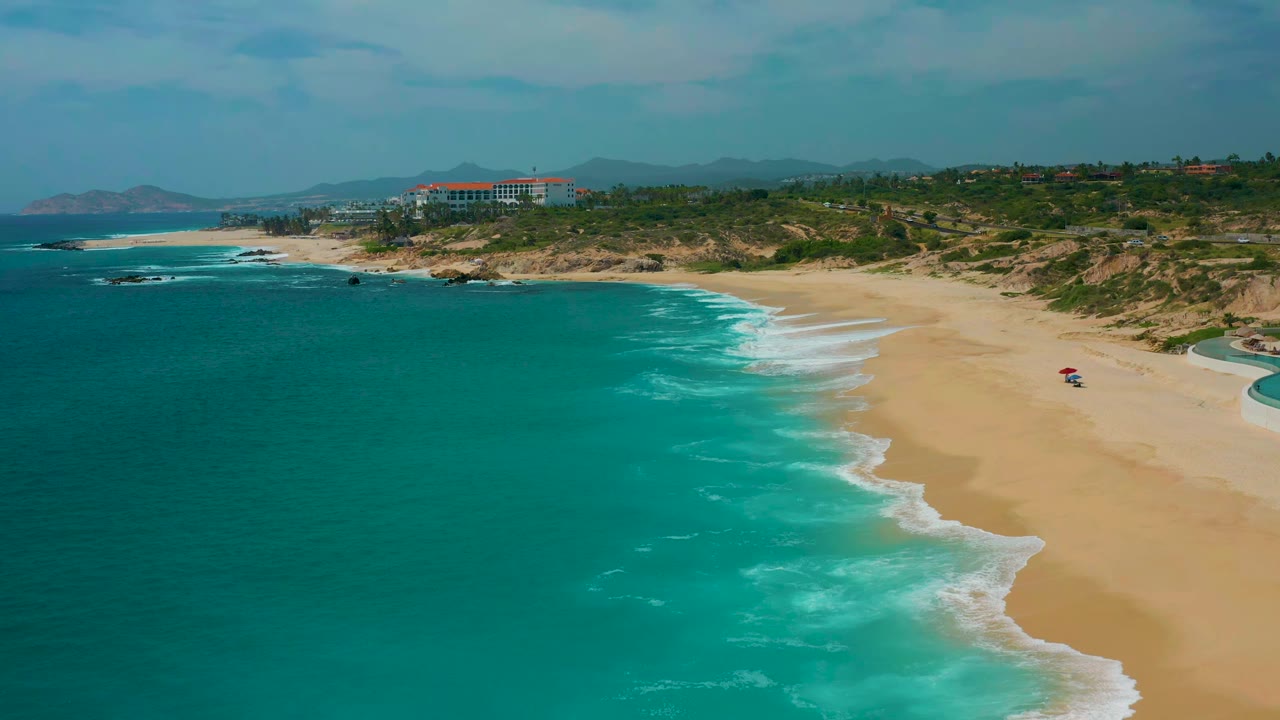 Aerial view over the Secrets Marquis Los Cabos, Mexico shoreline, with turquoise water on the left, surf bands along the sand, a red umbrella on the beach, and no visible people.
