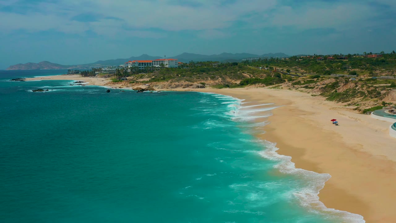 Aerial frame from Secrets Marquis Los Cabos, Mexico, showing open teal water, foamy waves reaching the beach, dune vegetation inland, distant hills, and no visible people.