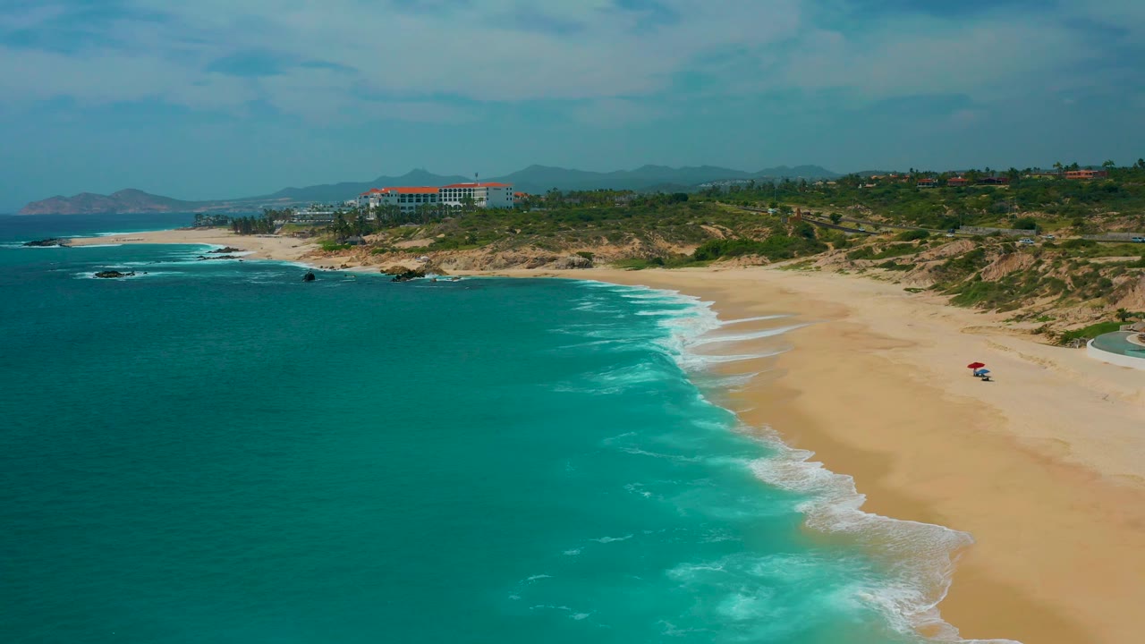 Aerial view near Secrets Marquis Los Cabos, Mexico, with the sea filling the left foreground, surf tracing the beach, dunes and buildings beyond the sand, and no visible people.