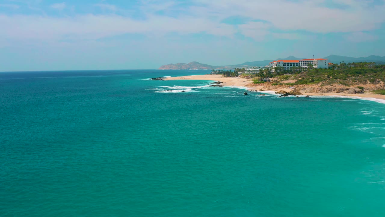 Aerial frame at Secrets Marquis Los Cabos, Mexico, showing more open turquoise water, a pale beach curving along the coast, white surf on rocks, resort buildings, and no visible people.