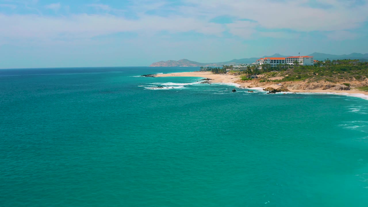 Aerial view at Secrets Marquis Los Cabos, Mexico, with blue-green water dominating the foreground, the beach and breaking surf set farther back, resort buildings to the right, and no visible people.