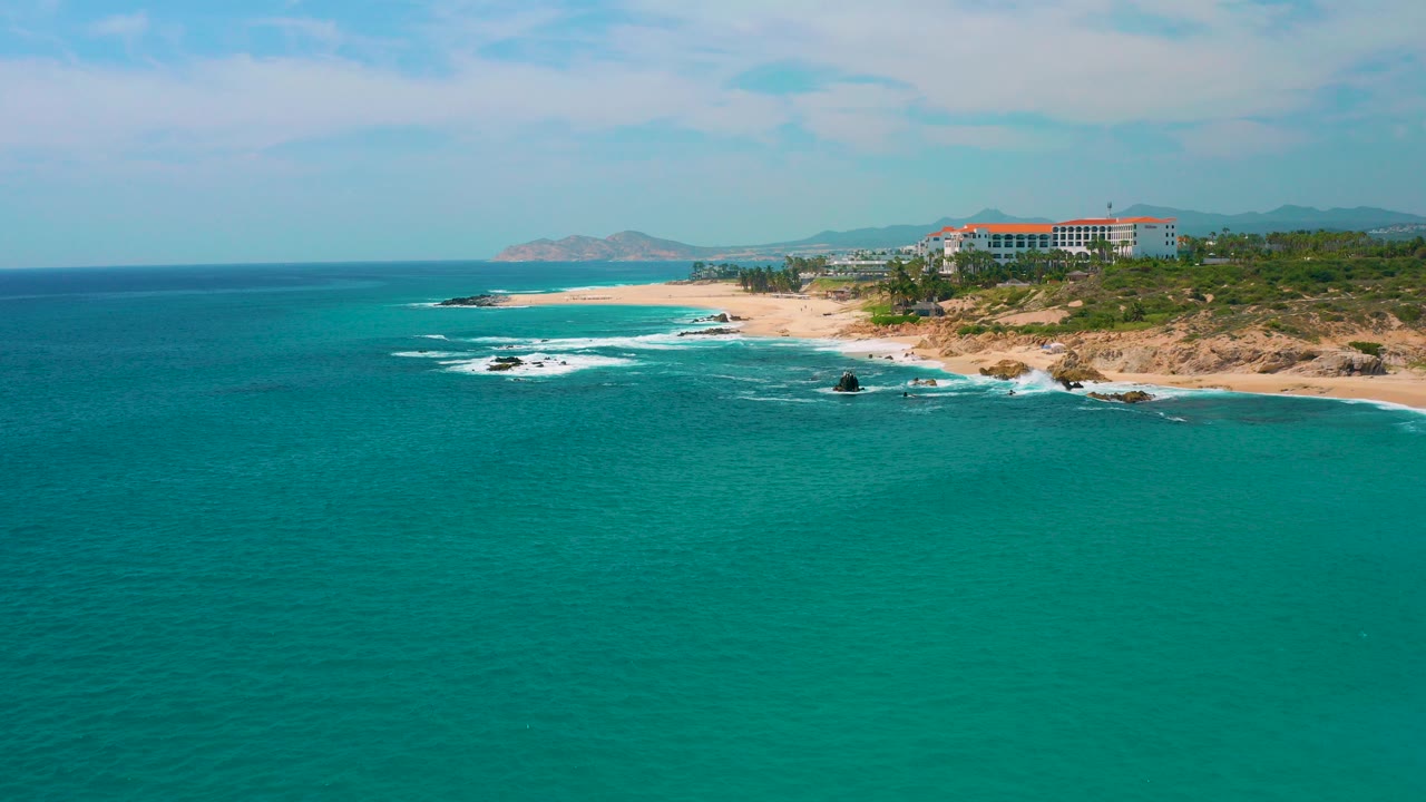 Wide aerial view at Secrets Marquis Los Cabos, Mexico, with turquoise water in the foreground, a sandy beach and rocky shoreline beyond, distant red-roofed buildings, and no visible people.