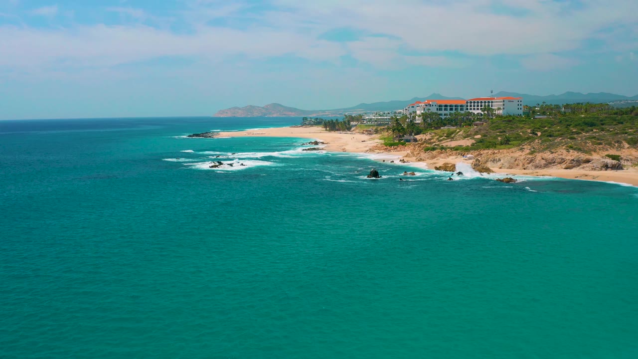 Drone-height coastal frame at Secrets Marquis Los Cabos, Mexico, with broad turquoise water below, a curved sandy beach, surf on rocks near shore, resort-area buildings on the bluff, and no visible people.