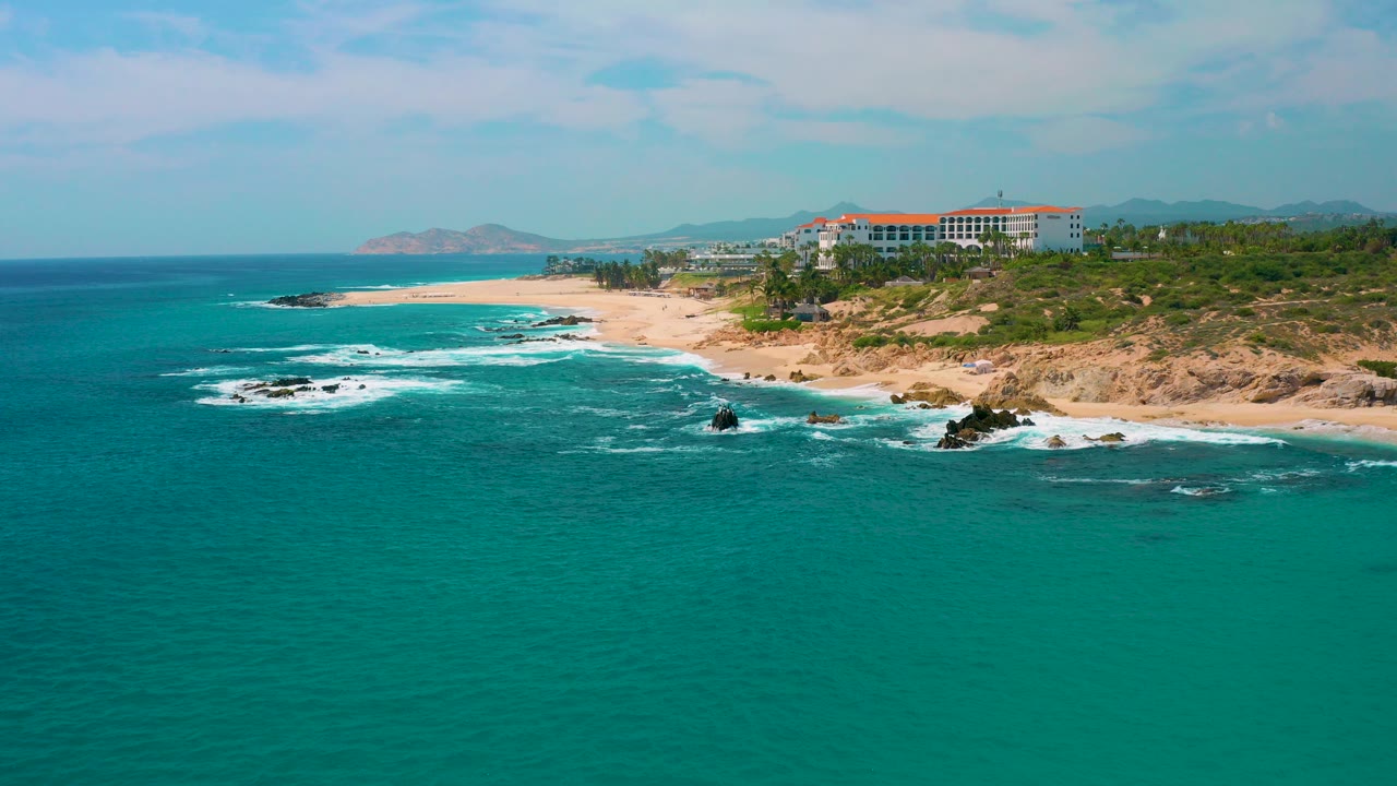 At Secrets Marquis Los Cabos, Mexico, turquoise ocean spreads across the foreground while white surf reaches rocks and a sandy beach below clifftop resort buildings, with no visible people.