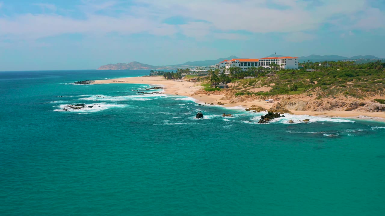 At 0:01, the aerial frame holds the Secrets Marquis Los Cabos resort near the upper right as surf bands curve along the beach and dark rocks break the turquoise water, with no visible people.