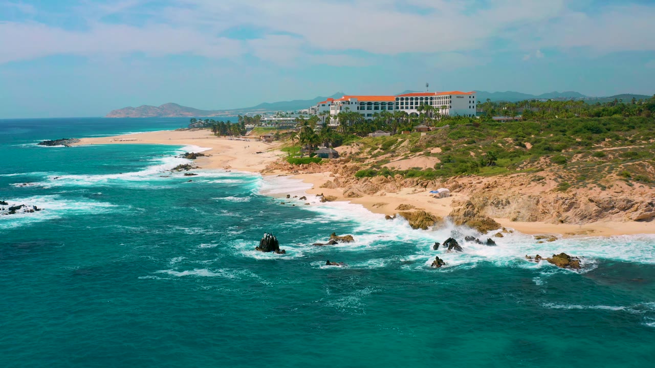 Aerial frame over Secrets Marquis Los Cabos, Mexico, showing more broken whitewater around offshore rocks, tan beach along the right side, the resort above the shore, and no visible people.