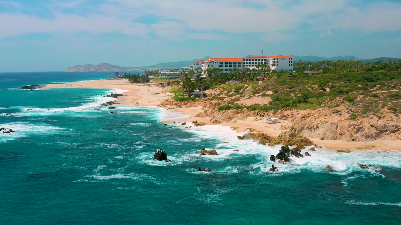 Aerial view at Secrets Marquis Los Cabos, Mexico, with the surf line pushed farther across the lower right rocks, open ocean to the left, the red-roofed resort in the background, and no visible people.