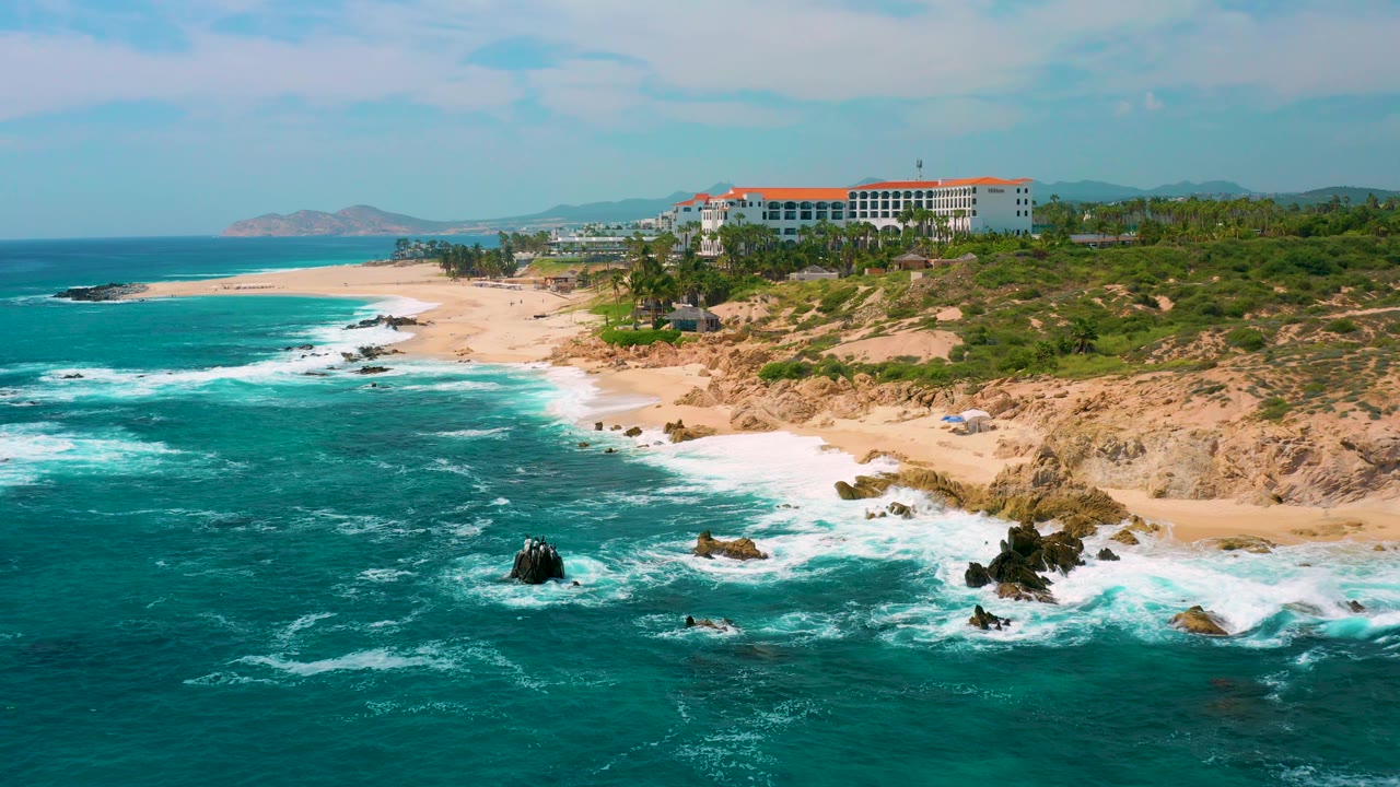 Mid-clip aerial frame at Secrets Marquis Los Cabos, Mexico, with a wider sweep of sandy beach, turquoise water, scattered rocks, coastal vegetation, the resort set back from shore, and no visible people.