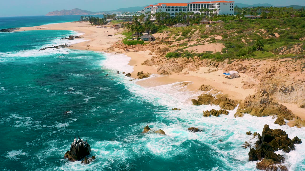 Aerial view at Secrets Marquis Los Cabos, Mexico, showing turquoise ocean, white surf, pale sand, rocky shoreline, distant resort buildings, and no visible people.