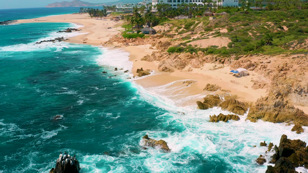 Aerial view at Secrets Marquis Los Cabos, Mexico, looking along a curved sandy beach where white surf wraps around rocks and resort buildings sit above the dunes, no visible people.