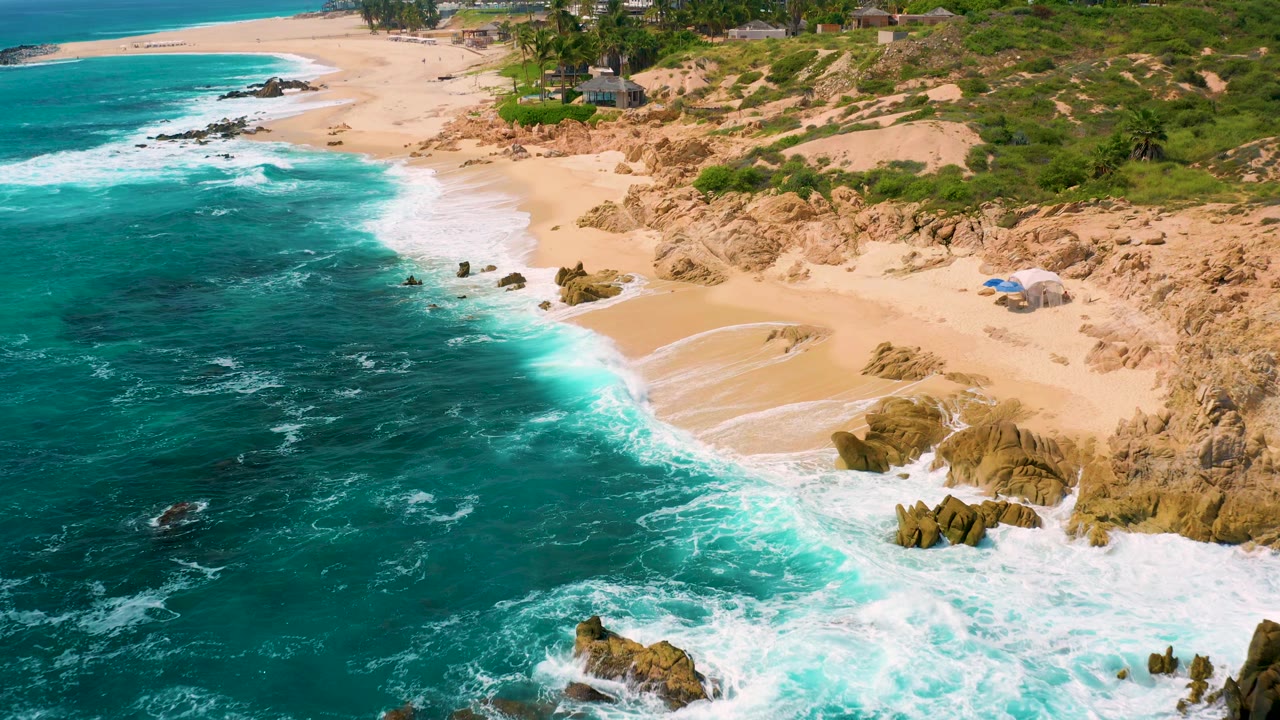 Closer aerial frame at Secrets Marquis Los Cabos, Mexico, with turquoise water on the left, white wash over pale sand, rocky outcrops on the right, and no visible people.