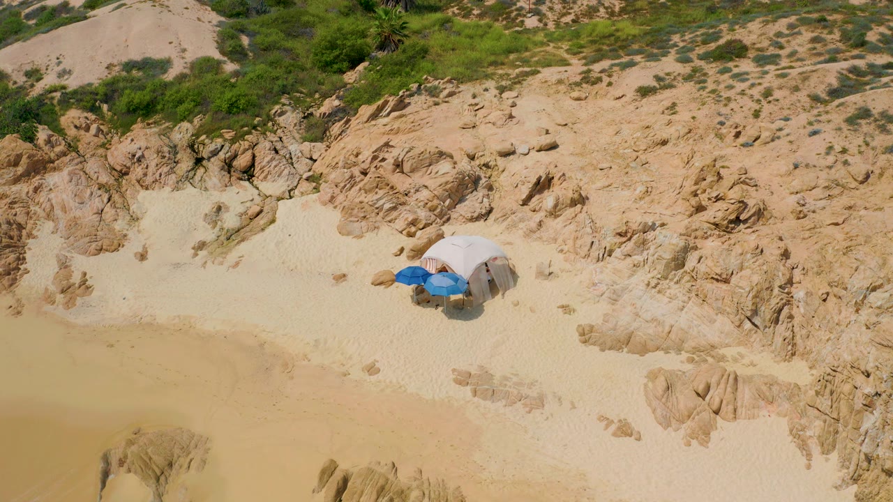 Overhead frame from Secrets Marquis Los Cabos, Mexico showing a small white shelter and blue beach gear on pale sand beside tan rocks and shallow water.