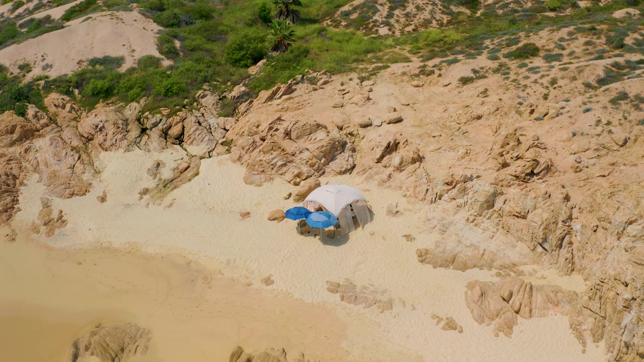 High overhead frame from Secrets Marquis Los Cabos, Mexico showing the beach shelter near the right side of the sandy pocket with rocks and scrub behind it.