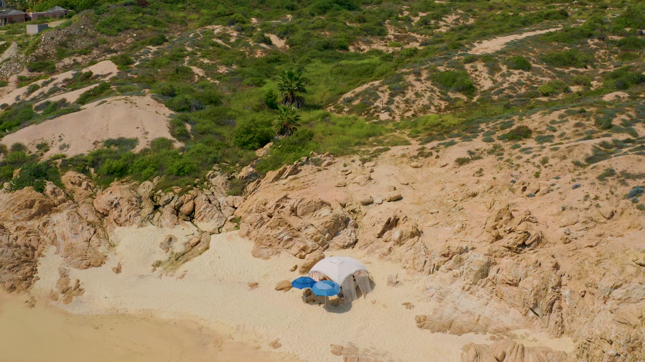 Aerial view of sandy rocks, green brush, and a blue umbrella near a beach shade.