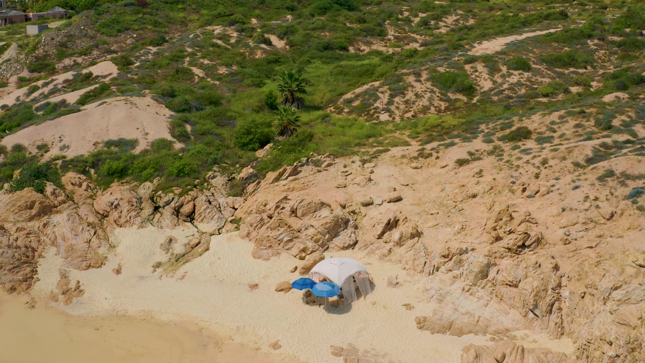 Aerial view at Secrets Marquis Los Cabos with the blue umbrella low in frame, rocky sand across the foreground, and dense green brush covering the slope, with no visible people.