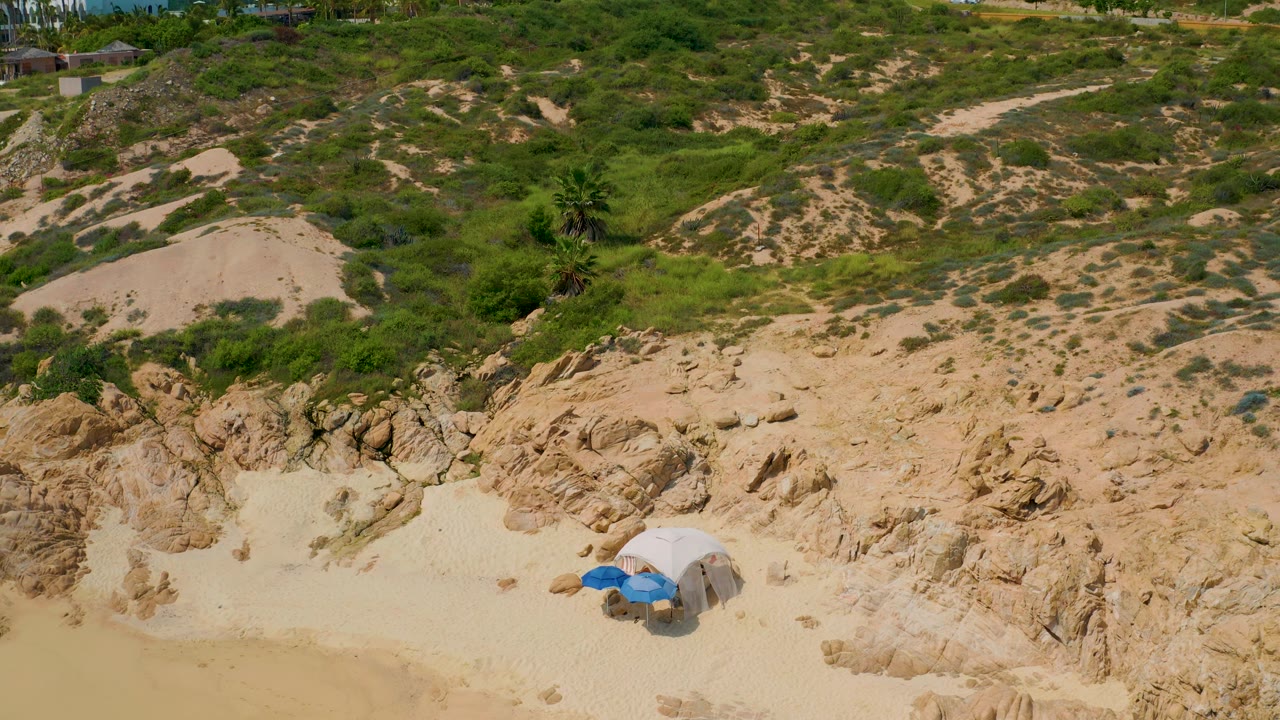 Aerial frame at Secrets Marquis Los Cabos showing the umbrella and shade structure near the bottom, tan rocks in the center, and green scrub across the upper hillside, with no visible people.