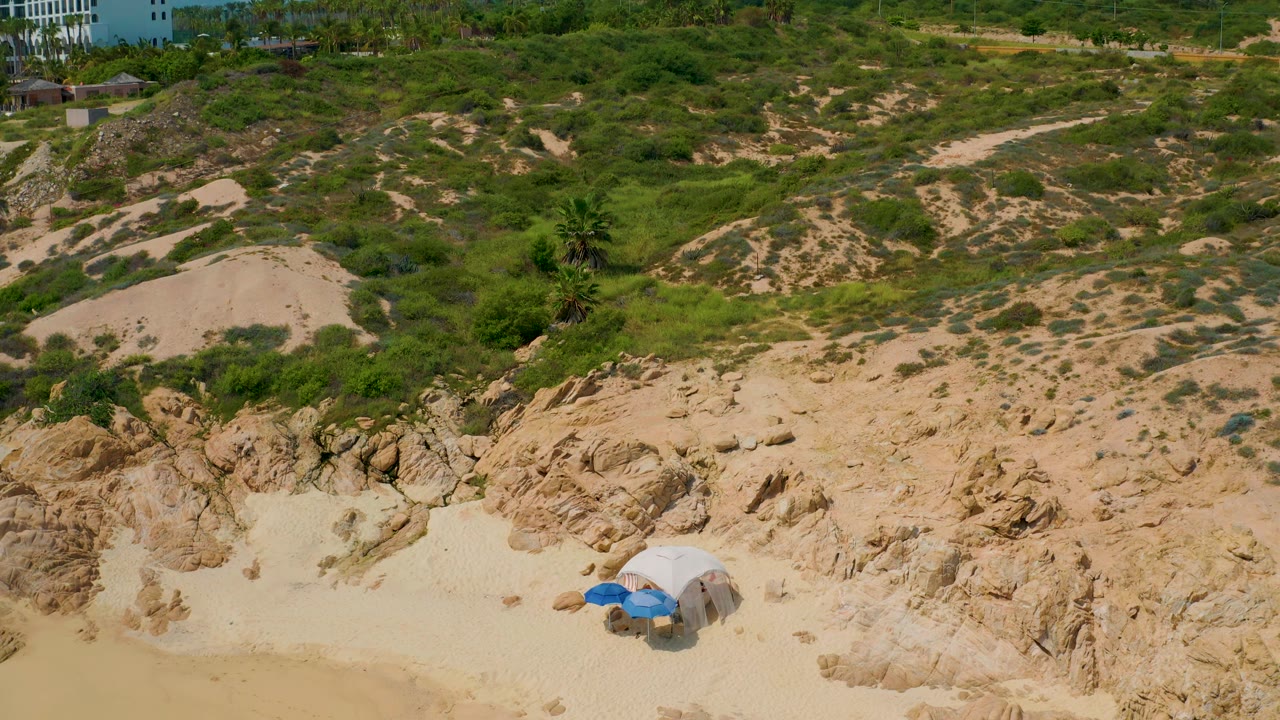 Aerial view at Secrets Marquis Los Cabos where white resort buildings enter the upper background above the scrubby hillside and sandy rock foreground, with no visible people.