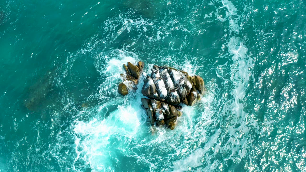 Overhead view of turquoise water at Secrets Marquis Los Cabos, Mexico, with white foam breaking around a compact dark rock cluster and no visible people.