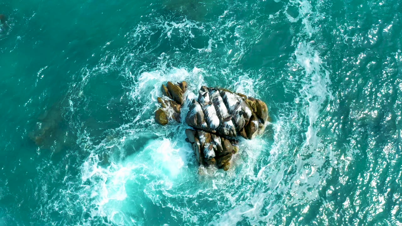Overhead shot at Secrets Marquis Los Cabos, Mexico, with blue-green water, bright foam curling around the right and lower edges of the rocks, and no visible people.