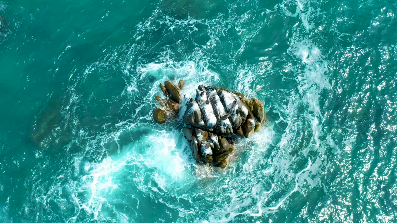 Near-vertical aerial view of Secrets Marquis Los Cabos sea, showing foamy water spreading below the central rocks while the upper water stays darker and no visible people are present.
