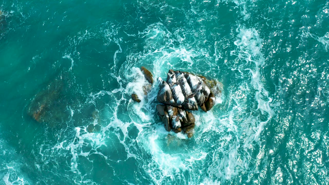 Opening overhead view at Secrets Marquis Los Cabos, Mexico, showing turquoise water and white surf circling a central dark rock cluster with no visible people.