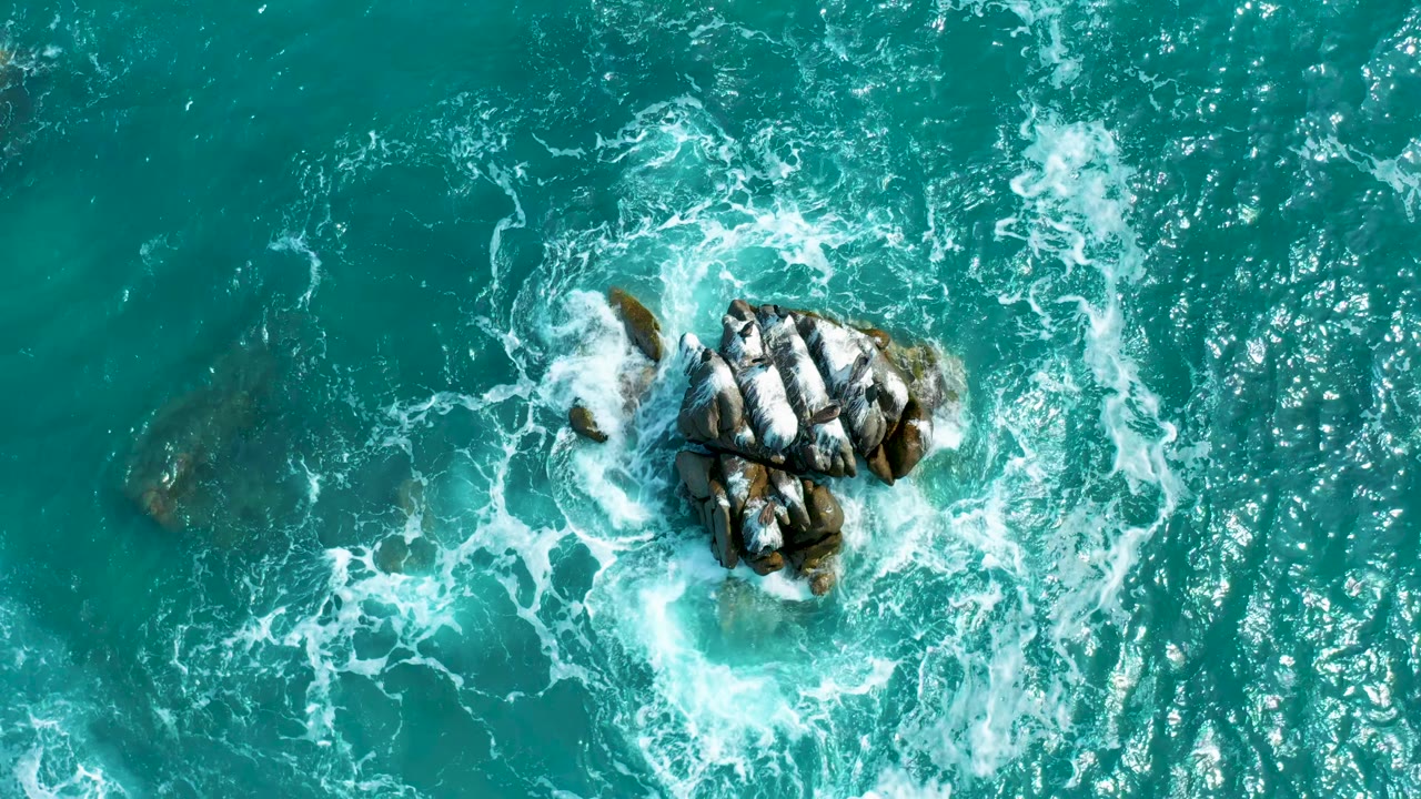 Whitewater curling around a central rock cluster in blue green ocean