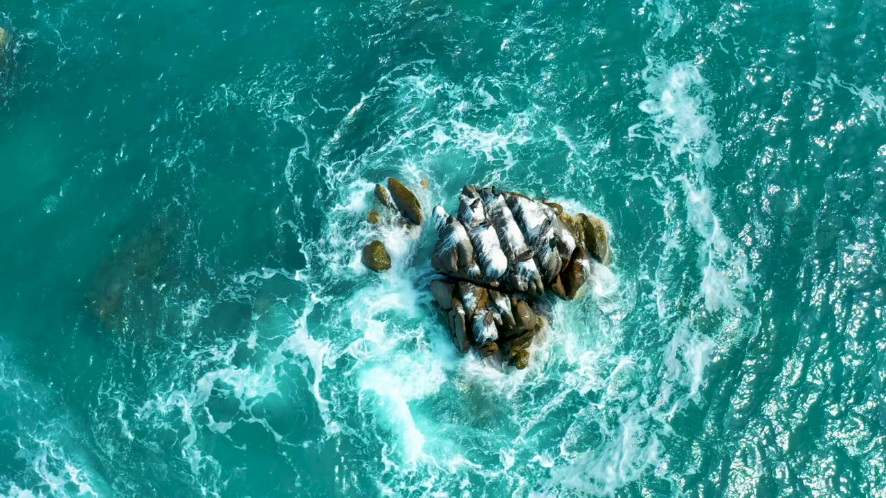 A top-down view at Secrets Marquis Los Cabos, Mexico, where the rocks sit slightly right of center and a brighter patch of foam trails below them, with no visible people.