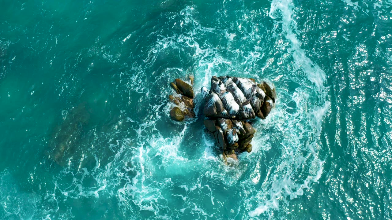 Overhead view near Secrets Marquis Los Cabos, Mexico, with turquoise water and white foam circling a dark rock cluster left of center, no visible people.