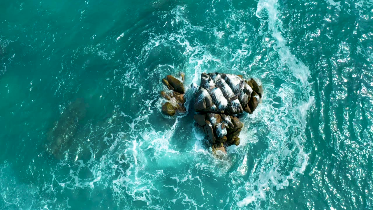 Aerial frame from Secrets Marquis Los Cabos, Mexico, showing white foam tightening around the central rocks with open turquoise water to the right, no visible people.