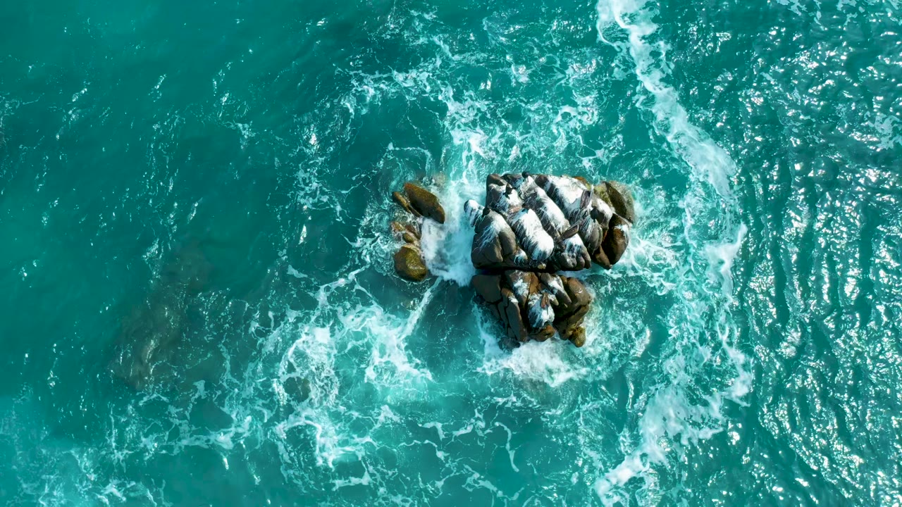 Top-down coastal view near Secrets Marquis Los Cabos, Mexico, with the rock outcrop slightly high in frame and white water spreading across the upper right, no visible people.