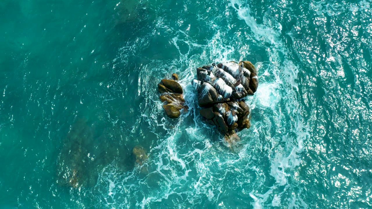 Overhead frame from Secrets Marquis Los Cabos, Mexico showing turquoise sea, white foam, a central rock cluster, faint submerged rocks, and no visible people.