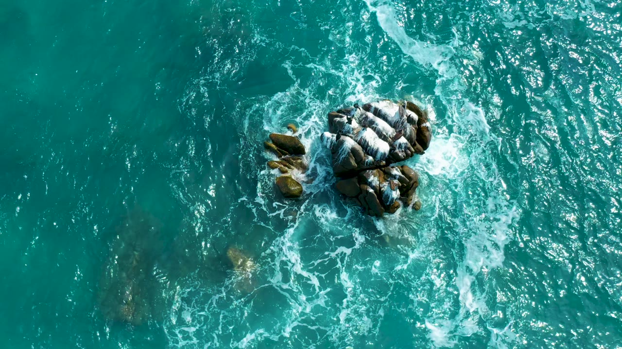 Top-down Secrets Marquis Los Cabos, Mexico frame with rocks shifted toward the upper right, churning white foam, rippled turquoise water, and no visible people.