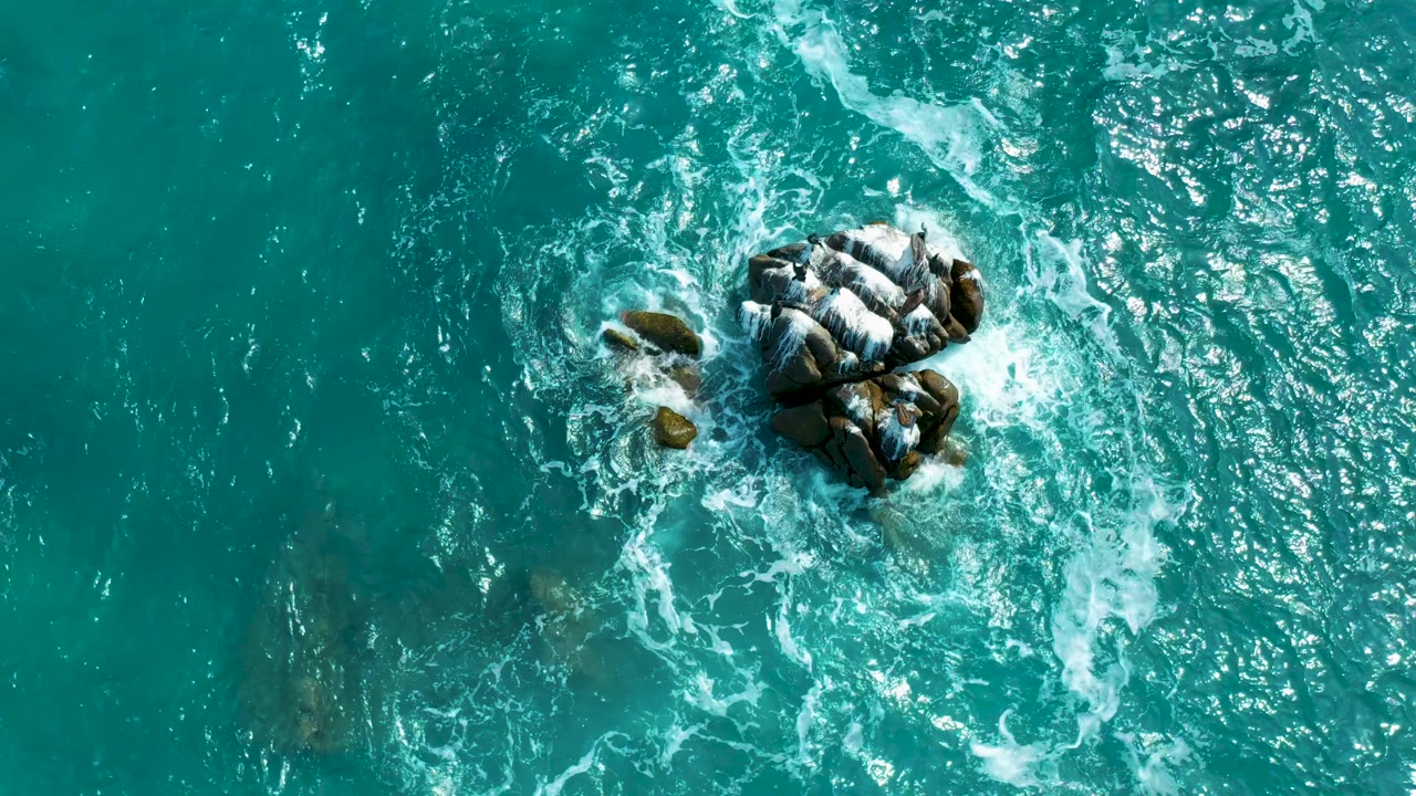 Overhead Secrets Marquis Los Cabos, Mexico shot with the rock cluster right of center, foam trailing across the turquoise water, darker submerged shapes, and no visible people.