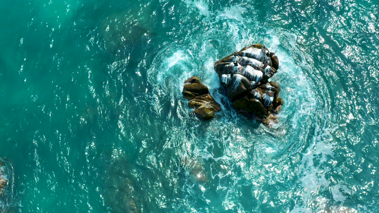 Top-down view of turquoise water swirling around dark rock formations at Secrets Marquis Los Cabos, Mexico, with bright foam and no visible people.