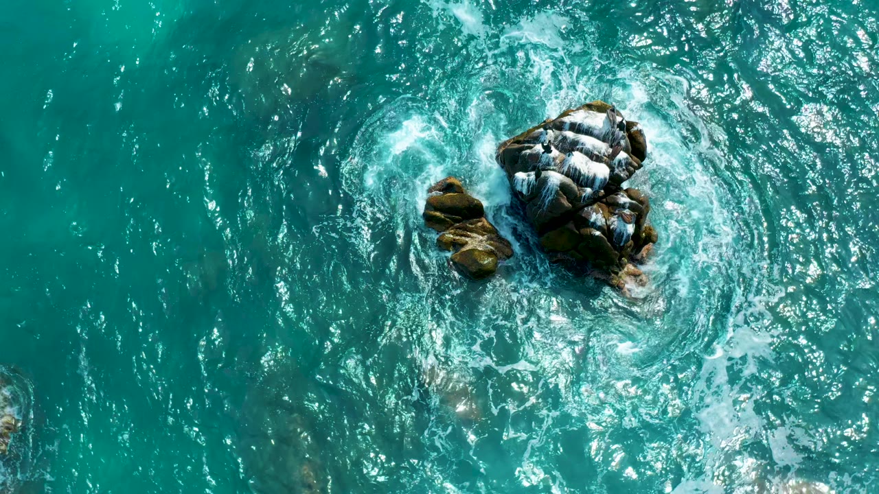 Aerial frame of a central dark rock cluster in turquoise water at Secrets Marquis Los Cabos, Mexico, with a broad white foam ring and no visible people.