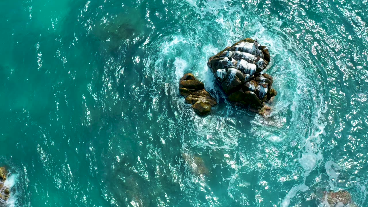 Blue-green ocean surrounds a rocky outcrop at Secrets Marquis Los Cabos, Mexico, as white surf breaks along the right side with no visible people.