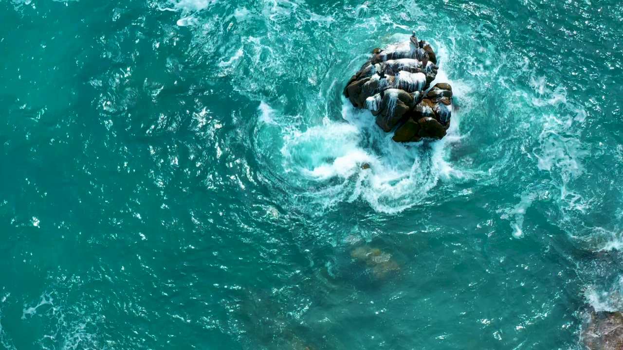 Overhead drone view at Secrets Marquis Los Cabos, Mexico, with white foam curling around the main exposed rocks while a faint lower rock remains visible below the surface, with no visible people.